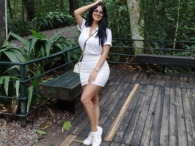 JennaDerose com uma camisa branca molhada e transparente, que revela pequenos seios e mamilos. Sua mão toca a parede, ela tem cabelo escuro, brincos de ouro e uma tatuagem no braço direito..
