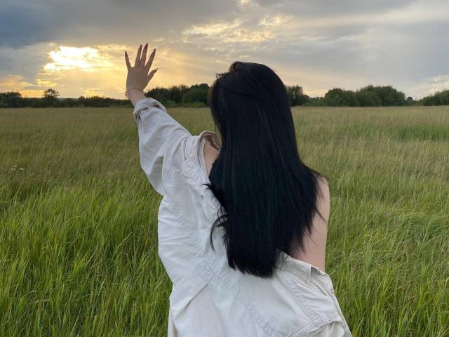 MertieDasinger with dark hair, wearing a light blue denim jacket and black shirt, holding the jacket's hood, standing on grassy field, under cloudy sunset.