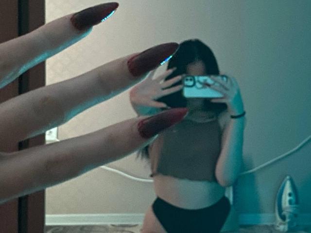 MertieDasinger taking a selfie in a mirror, wearing a black sports bra, with hands covering her mouth, phone in front, hair in a bun, in a dimly lit room.