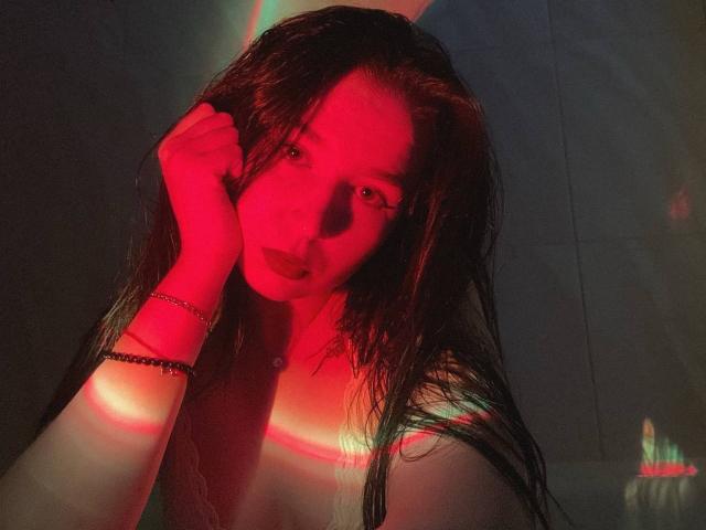 MertieDasinger with long black hair, wearing a black spaghetti strap top and red skirt, head tilted back, eyes closed, purple light, silver hair clip, sitting on a bed.