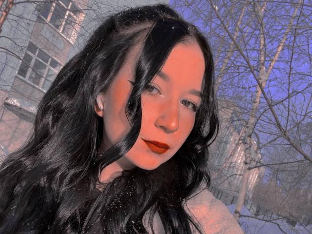 MertieDasinger with long black hair, red bow, beige jacket, black bra, holding phone, mirror selfie, showing tits, holding hair with left hand, glitter phone case.