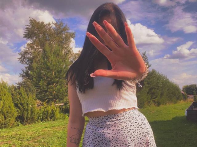 MertieDasinger with long brown hair, white crop top, and white patterned high-waisted pants, raising her hand in front of her face, showing a tattoo on her arm.