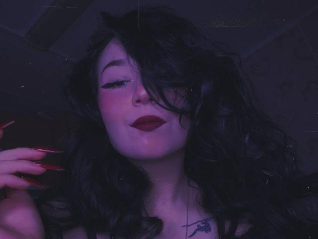 MertieDasinger has long black wavy hair, closed eyes, and red lipstick. She's wearing a white glittery jacket, standing in snow with a blue-purple light on her face.