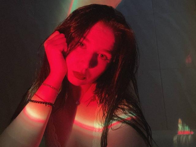 MertieDasinger is leaning back, eyes closed, with long dark hair, wearing a loose white shirt and dark pants, her hand on the floor, head tilted up.