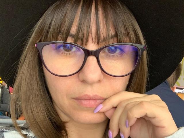 CrissyLove met bruin haar, een zwarte hoed, paars getinte brillen en paarse nagels, die een zuur gezicht trekt met haar vinger op haar lip, en haar lelijke gezicht laat zien..