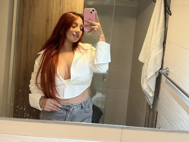 CereScarlett with long red hair, white unbuttoned shirt showing her tits, gray pants, holding pink phone, smiling in mirror, bathroom, white towel on right.