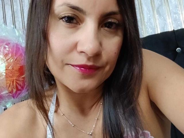 SolaraDubois has brown eyes, straight dark hair, pink glossy lipstick, a small beauty mark under her right eye, and wears a silver necklace with a chain. Boobs visible.