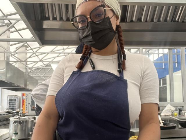GalaMalkova with glasses, black mask, white shirt, blue apron, braided red hair, white headscarf, medium tits, standing in a kitchen, metal ceiling, stainless steel appliances.