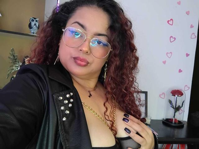 SophiLarusso with curly red hair, blue-tinted glasses, black leather jacket, gold necklace, and black mesh top, showing her tits and pink lips, with black nail polish.