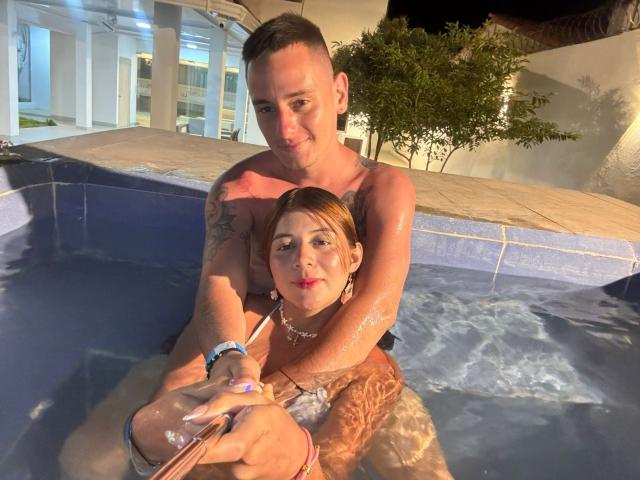 MeganDante, met kort bruin haar en kleine borsten, zit in een blauw betegelde jacuzzi, met een drankje in haar hand, en wordt van achter vastgehouden door een gespierde man zonder shirt, die tatoeages heeft..