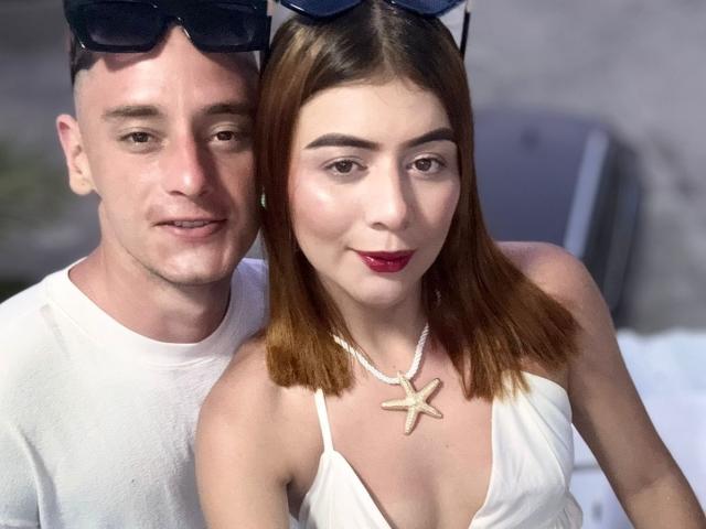 MeganDante mit braunen Haaren, einem weißen Kleid, einer Sternkette und rotem Lippenstift hat einen Mann mit weißem Hemd, der eine Sonnenbrille auf dem Kopf trägt, der hinter ihr sitzt..