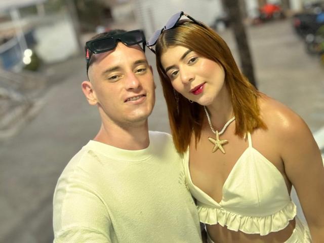 MeganDante mit hellbraunen Haaren, die ein weißes Bikini-Oberteil und eine Sternkette trägt, steht neben einem kahlköpfigen Mann in einem weißen Hemd, beide lächeln. Ihre Brüste sind sichtbar..