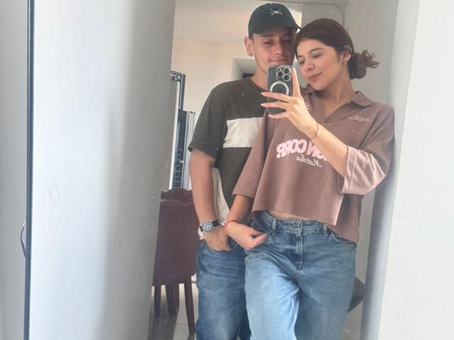 MeganDante, avec des cheveux bruns attachés en queue de cheval, portant une chemise rose et un jean bleu, se tient à côté d'un garçon en chemise verte et blanche, et prend un selfie devant un miroir..