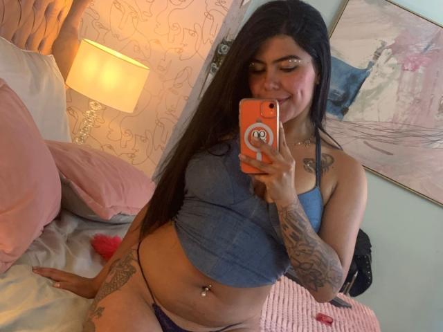 CatalinaReiyna with long dark hair, blue crop top, tattoos on arm and thigh, purple thong, holding orange phone, taking mirror selfie, smiling, pink pillows, bed, lamp, painting.