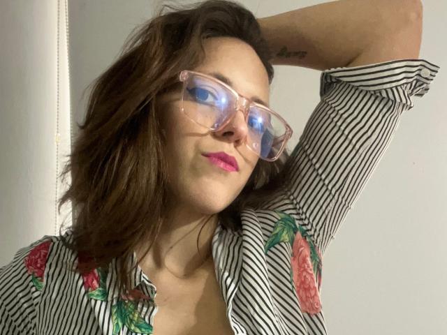 LunaFiorella, avec son rouge à lèvres rose, sa chemise rayée, ses motifs floraux et ses lunettes transparentes, a le bras levé, montrant un tatouage, et a les cheveux bruns et ondulés. Ses seins sont partiellement visibles..