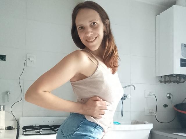 LunaFiorella mit roten Haaren, lächelnd, trägt ein weißes Tanktop und blaue Jeans, eine Hand am Hüfte, zeigt ihr Gesäß, steht in einer Küche..