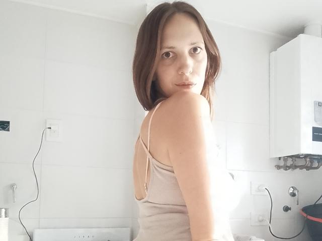 LunaFiorella, con i capelli castano di lunghezza media, indossa una canotta beige, guarda oltre la spalla, mostrando la schiena nuda e piccoli seni, in una cucina con piastrelle bianche..