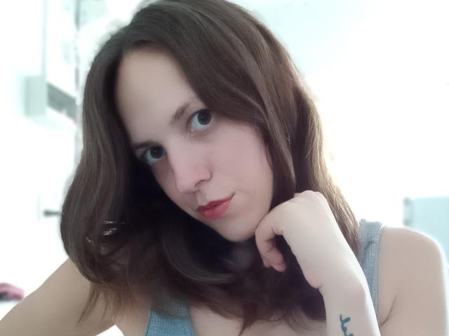 LunaFiorella with brown wavy hair, blue eyes, nose ring, and grey tank top, resting chin on hand, showing tattoo on wrist. Soft light, slightly blurred background.