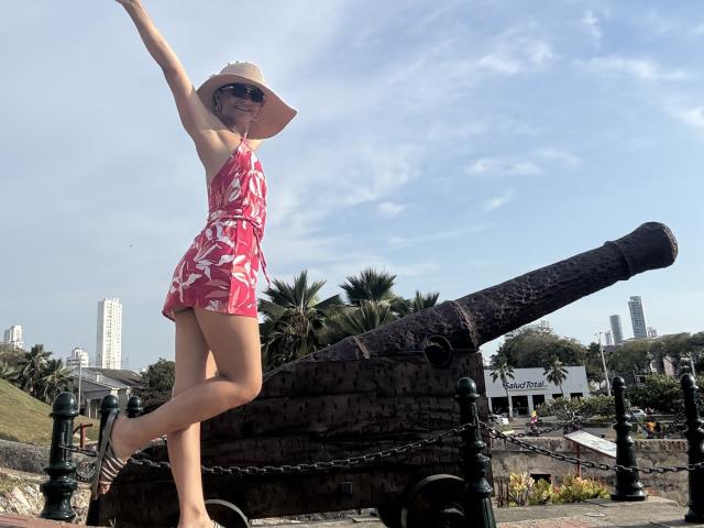KimmyK with sunhat, red floral dress, and sunglasses, arms raised, standing on cannon, showing her ass, palm trees, and city skyline in background, wearing striped shoes.
