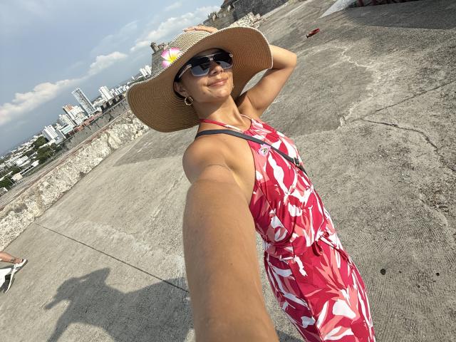 KimmyK with tan skin, wearing a red floral dress, sun hat, and sunglasses, holding a phone for a selfie. Her tits are small, and she's smiling. Urban rooftop.