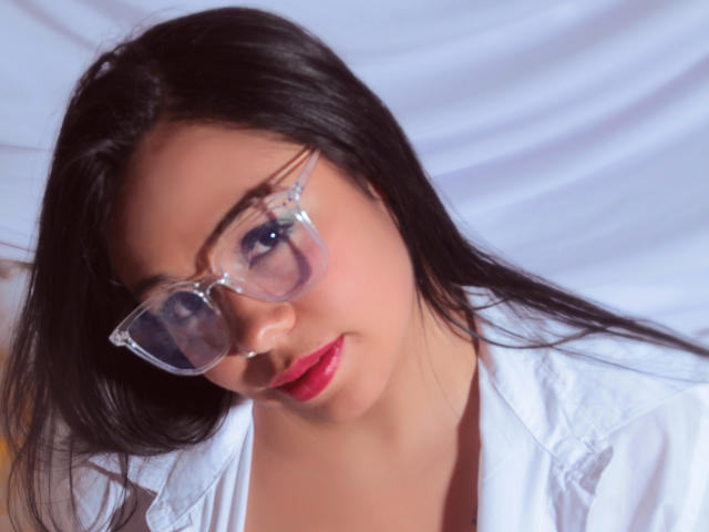 AmyNoa with black hair, wearing clear glasses and a white shirt, has red lips and a cute face, looking down with a slight tilt. Her ass is not visible.