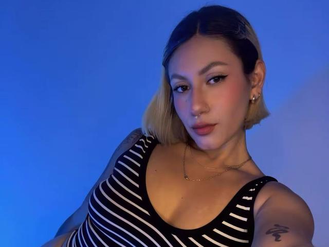 EmilyRobertts has blonde bob, black eyeliner, nose ring, and striped black-and-white top, showing small tits and tattoo on right arm, against blue background.
