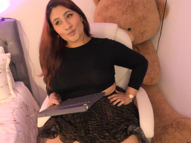 ElyRoss with brown hair, black top, and zebra-pattern skirt, sitting on white chair, holding laptop, huge tan teddy bear behind, wearing gold watch, white bed to left.