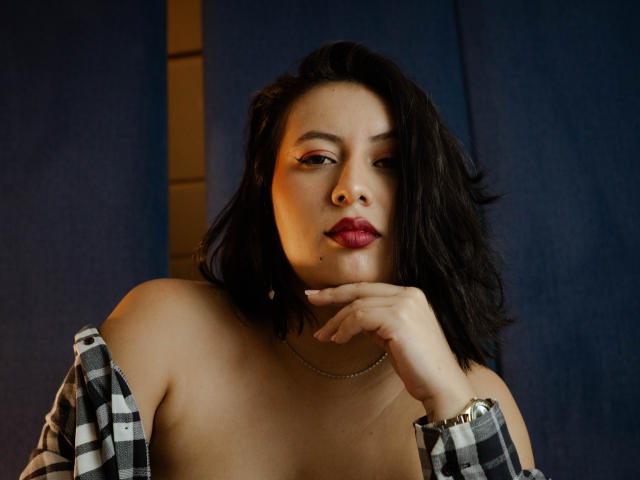 AmyRoss with dark wavy hair, pink eyeshadow, red lipstick, and a checkered shirt off her shoulders, touching her chin with a gold watch on her wrist.