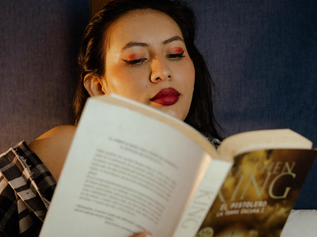 AmyRoss with red makeup, dark hair, and red lips, reading a book, her nose slightly upturned, focused, wearing a checkered shirt, book in front, slightly out of focus.