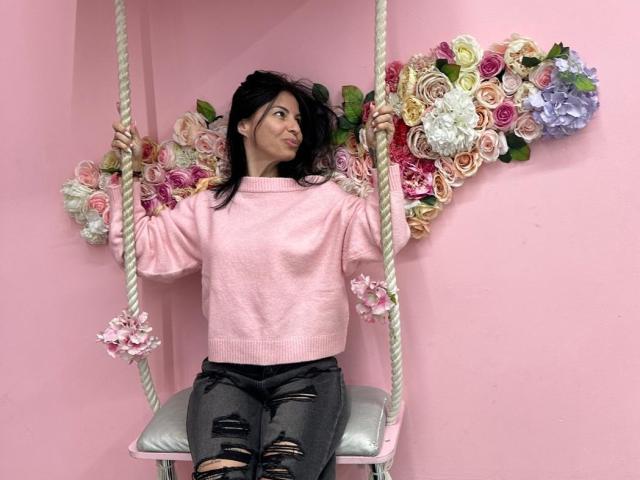 CrissyLove sitting on a pink, floral-decorated swing, wearing a pink sweater and ripped gray jeans, with black hair and a slight smile, looking to the side.