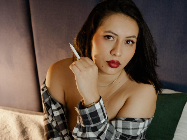 AmyRoss with dark hair, red lipstick, holding a pen to her ear, wearing an off-shoulder black-and-white checkered shirt, exposing her tits, and a necklace.