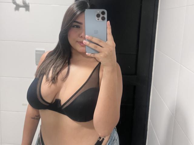 AlannaL with medium skin tone, long dark brown hair, wearing a black sheer bralette, showing her big tits, holding a silver iPhone in a bathroom mirror selfie.