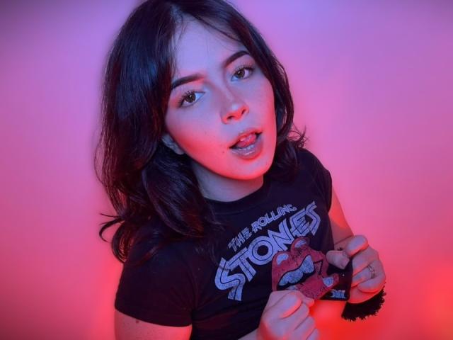 AliinaFontaine with wavy dark hair, wearing a black The Rolling Stones t-shirt, holding a ring with her fingers, mouth slightly open, pink and blue lighting on her face.