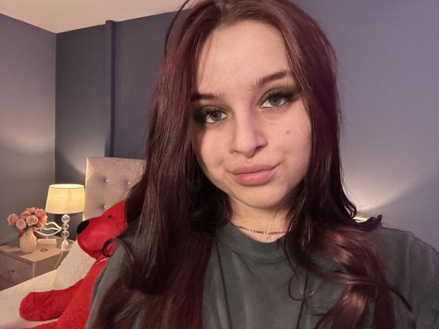 LucieRed has long, dark red hair, green eyes, and a slight pout. She's wearing a gray shirt, with a red pillow and lamp in the background.
