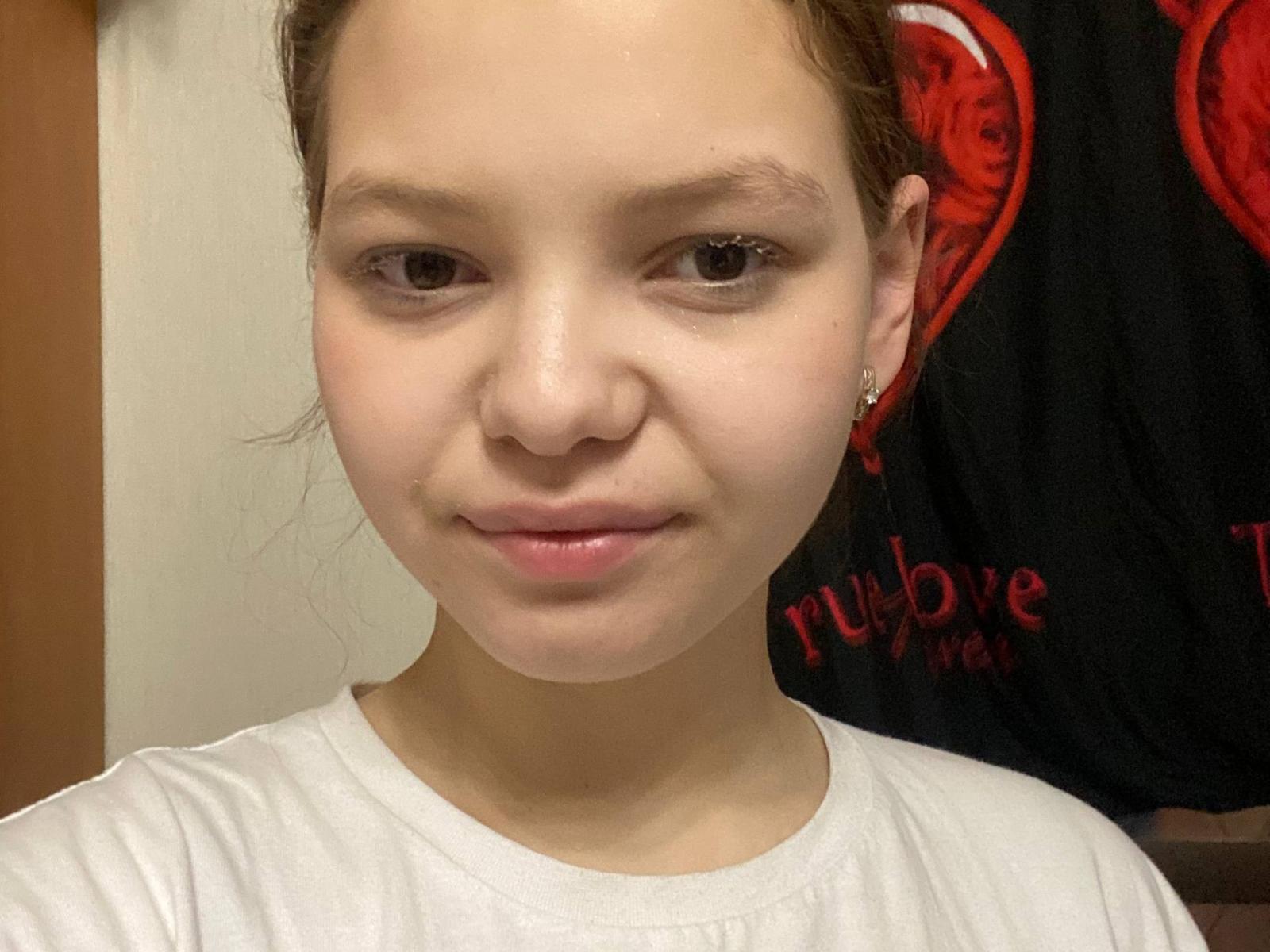Le visage d'ElmaAmaya, avec sa peau claire, ses yeux marrons et son petit nez, arbore un sourire discret. Elle porte une chemise blanche, ses cheveux bruns sont attachés en chignon, et elle a de petites boucles d'oreilles..