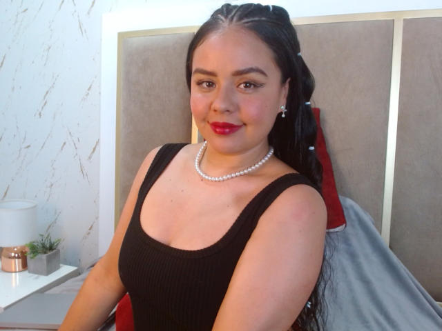 ArielleWilde with red lipstick, black ribbed tank top, pearl necklace, and long black braids, sitting on a bed, smiling. Boobs are visible, but not the focus.