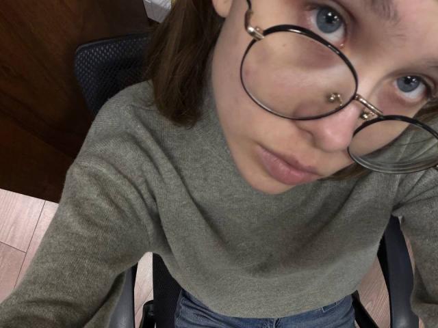 DorianArdis mit runder Brille, grauem Pullover und blauen Augen, blickt auf die Kamera, trägt einen Nasenring und sitzt auf einem Bürostuhl..