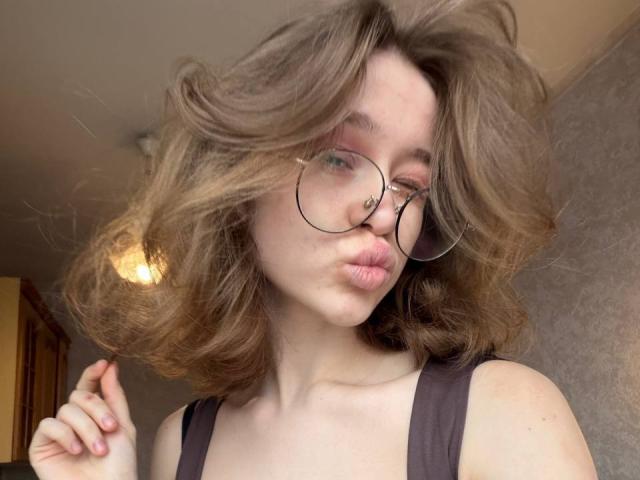 DorianArdis, con cabello castaño ondulado, labios carnosos y gafas, vestida con una camiseta oscura, soplando un beso, mostrando un piercing en la nariz y sujetando su cabello..