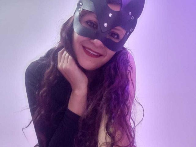 Arianna, avec de longs cheveux bruns ondulés, portant un masque de chat noir et souriant, touchant son cou, vêtue d'un haut noir, éclairage violet, mettant en valeur son rouge à lèvres et ses dents..