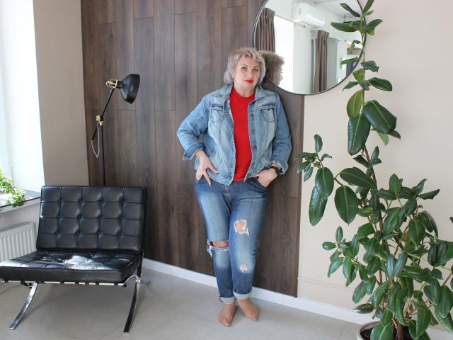 RenataDaster with blonde hair, red turtleneck, and ripped blue jeans, standing against a wooden wall, hands in pockets, next to a black chair and green plant.