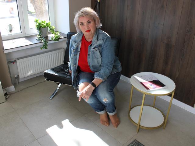 RenataDaster with short blonde hair, red shirt, denim jacket, and ripped jeans, sitting on black chair, holding cigarette, gold round table with book and phone, wooden wall.