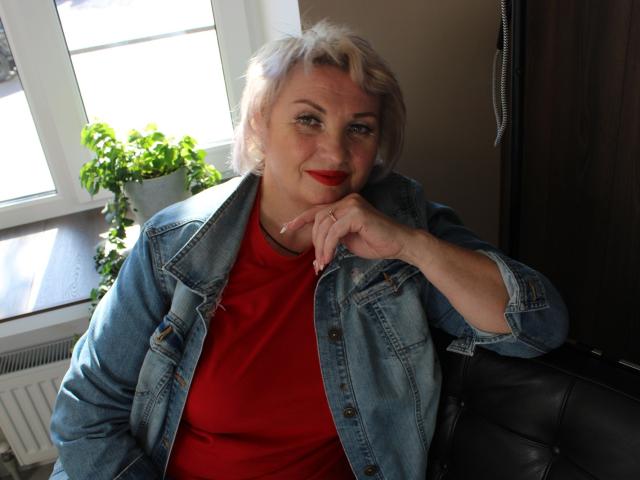 RenataDaster with short, grayish hair, red lipstick, denim jacket, and red shirt, resting chin on hand, sitting next to black leather chair, potted plant on window sill.