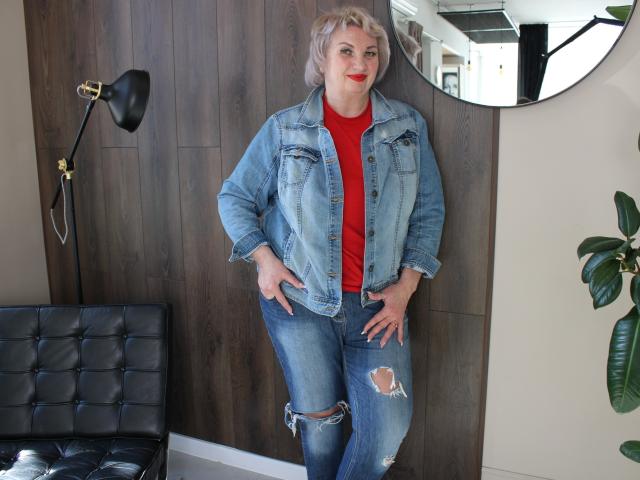 RenataDaster steht mit den Händen in den Hüften da, trägt ein rotes Hemd, eine blaue Jeansjacke und zerrissene Jeans, lächelt mit rotem Lippenstift, hat blondes Haar und eine kurvige, wohlgeformte Figur..