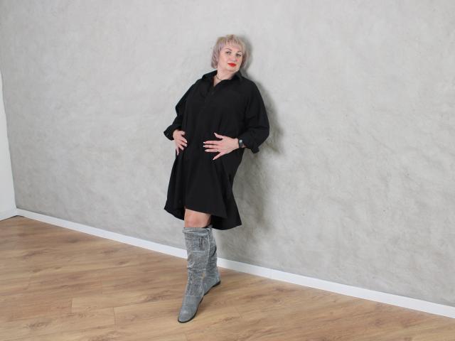 RenataDaster with short blonde hair, red lipstick, black dress, and gray knee-high boots, standing against a gray textured wall, hands on her belly, wooden floor.