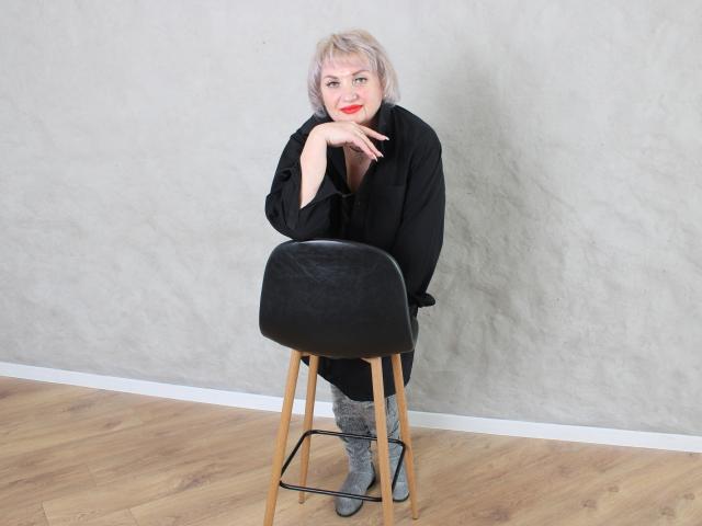 RenataDaster with pale blonde hair, red lipstick, black sweater, gray boots, and black stool, leaning on her hand, wooden floor, gray textured wall.