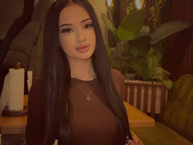 ShirleySiegel has long black hair, a cute face, full lips, and wears a black top with a gold necklace. She's standing in a warm, cozy indoor setting.