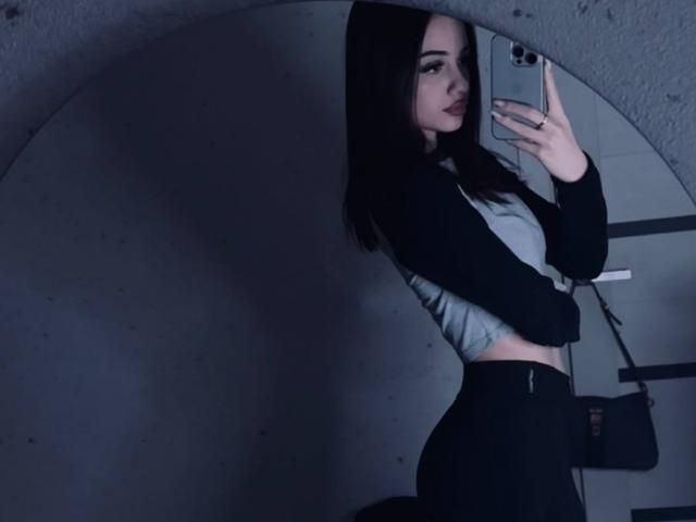 ShirleySiegel with long dark hair, wearing a black and gray top, black pants, taking a mirror selfie, showing her side with a slight curve to her ass.