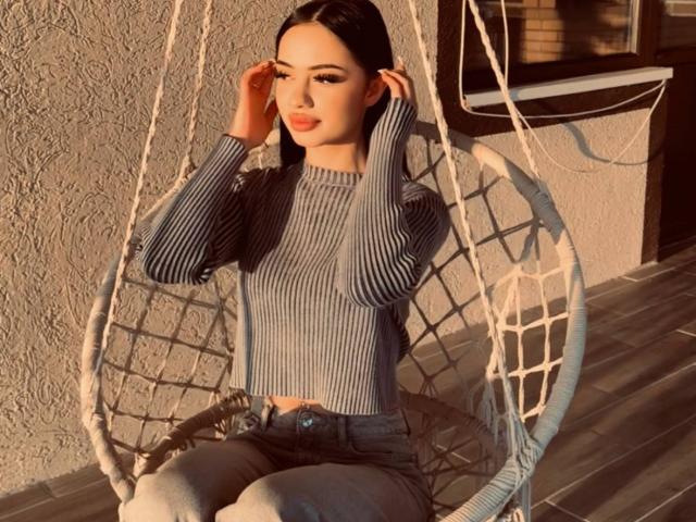 ShirleySiegel with dark hair, wearing a ribbed gray top, sitting in a hanging chair, hands in hair, blue jeans, soft sunlight, textured wall, wooden floor, relaxed, pretty.