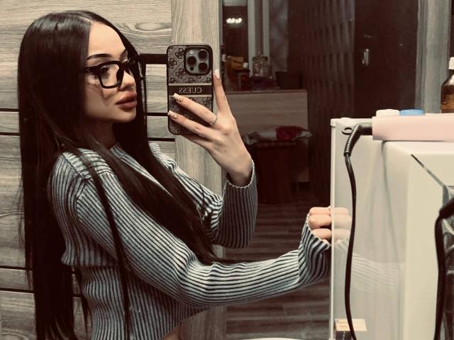ShirleySiegel with long black hair, wearing glasses and a striped shirt, taking a mirror selfie with her phone, showing her tits and holding a charger.