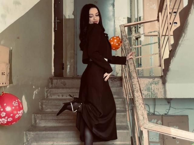 ShirleySiegel, with long black hair, wearing a black dress and high boots, standing on worn stairs, holding a red and orange balloon, looking over her shoulder.